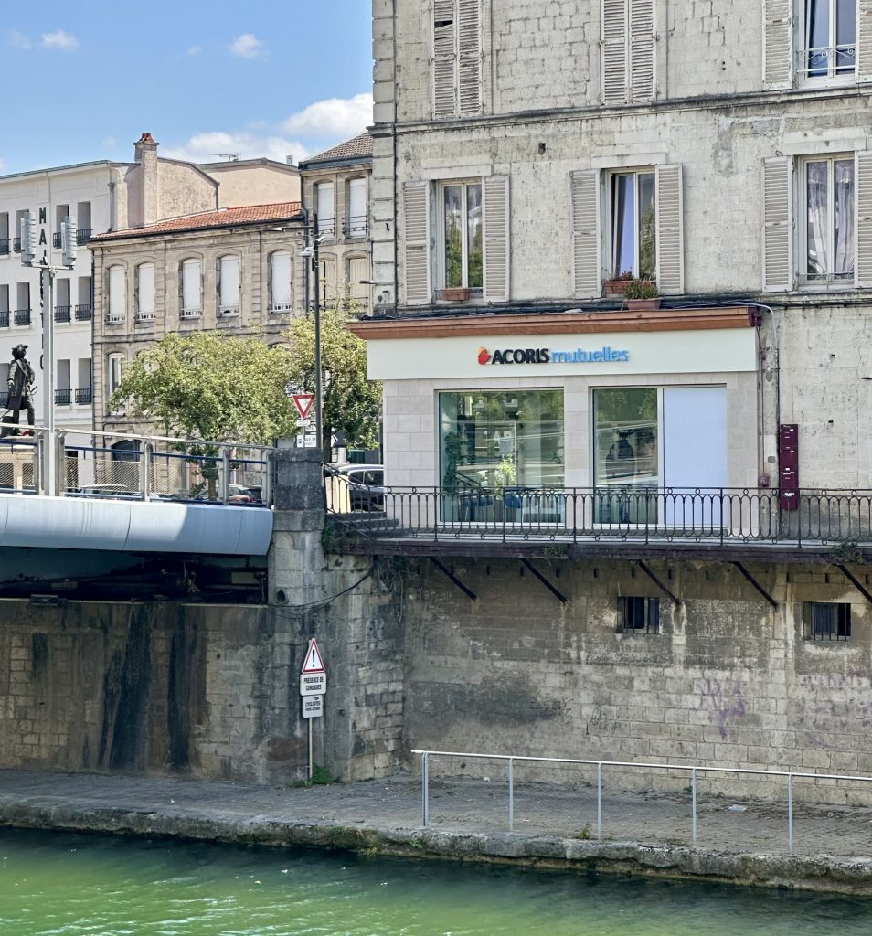 Façade d’une agence Acoris Mutuelles située au bord d’un canal, dans un bâtiment ancien en pierre aux volets blancs. Au premier plan, l’eau verte du canal longe un quai en pierre. À gauche, un pont métallique traverse le cours d’eau. Le ciel est bleu avec quelques nuages, et plusieurs immeubles urbains sont visibles en arrière-plan. Le logo « ACORIS Mutuelles » apparaît au-dessus de la vitrine de l’agence.