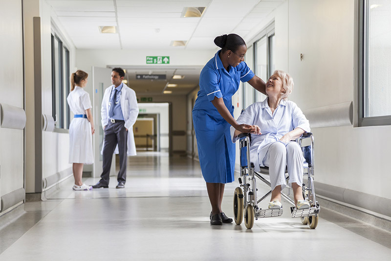 Infirmière aide une senior en fauteuil, médecins discutent, illustrant l'importance de la complémentaire santé.