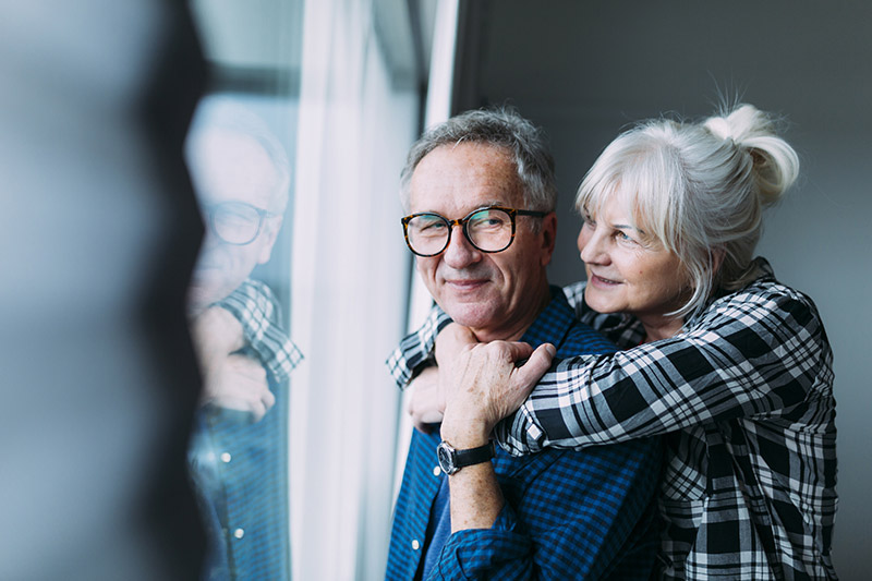 Couple senior souriant enlacé près d'une fenêtre, sérénité grâce à la complémentaire santé.