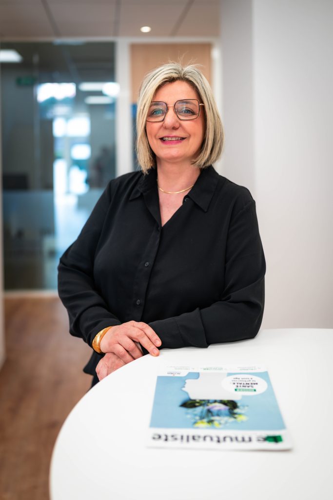 Femme souriante aux cheveux blonds accoudée à une table.