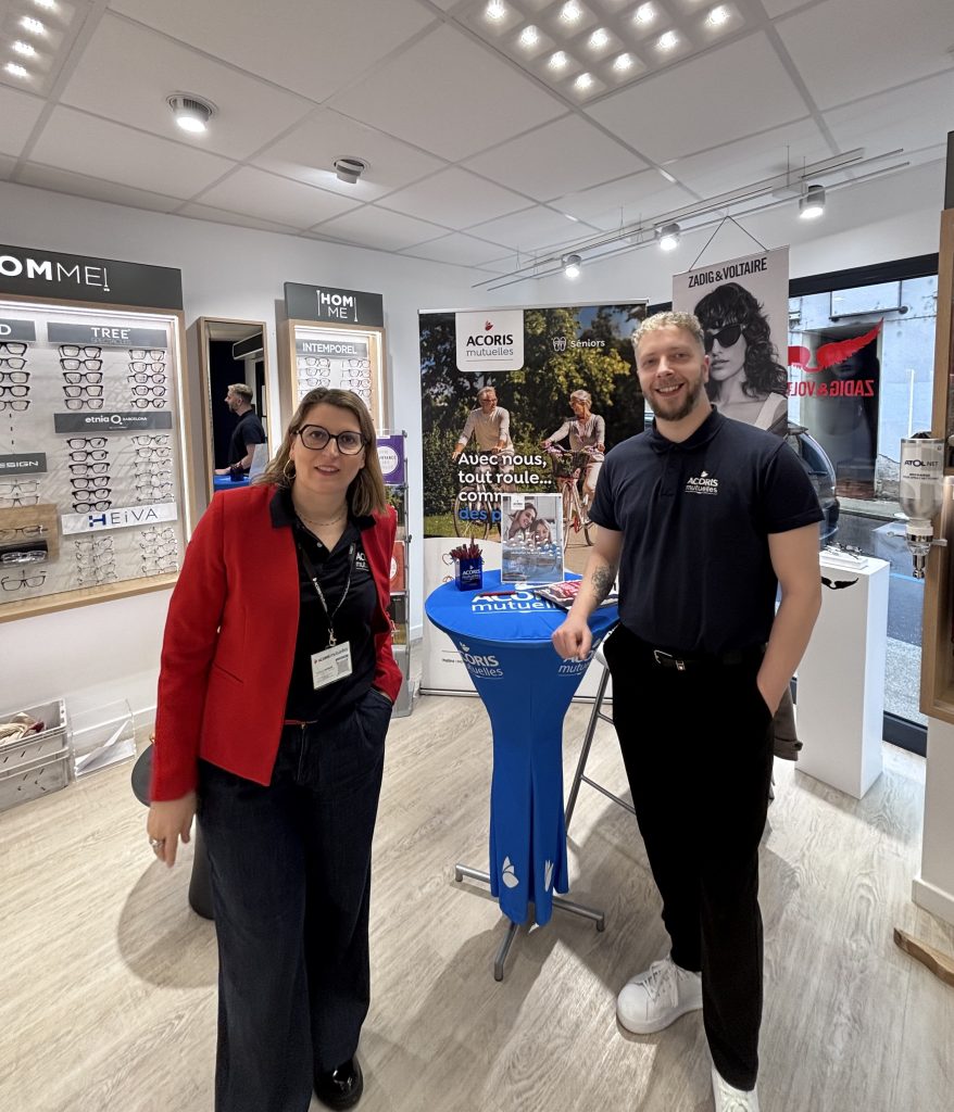 Deux personnes discutent mutuelle santé dans un magasin d'optique devant lunettes et affiches promotionnelles.
