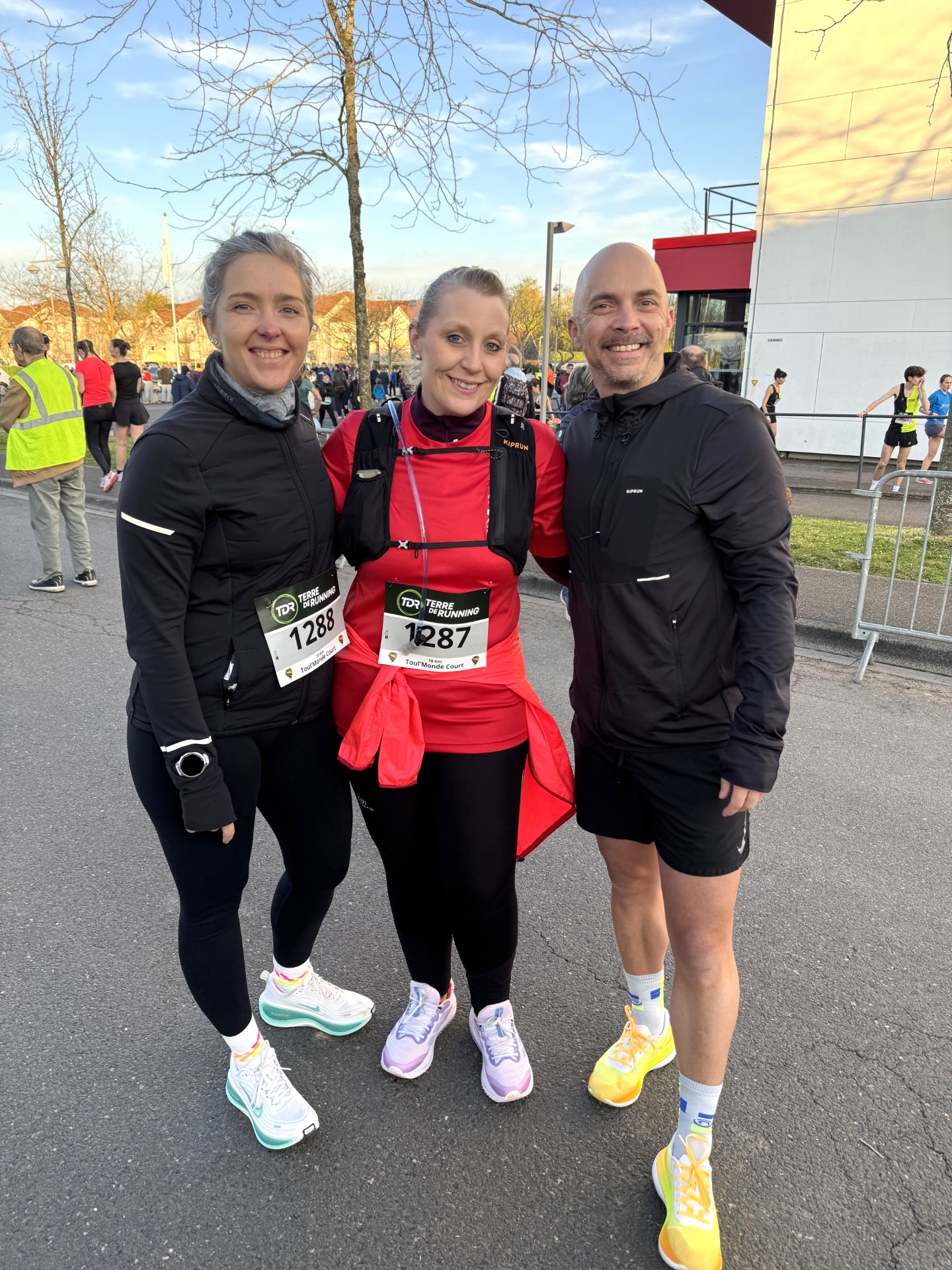 Trois personnes posent ensemble avant ou après une course à pied, portant des tenues sportives et des dossards, lors d’un événement sportif.