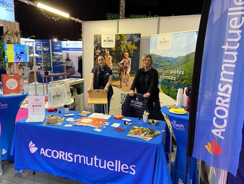 Stand ACORIS Mutuelles sur un salon, avec une table habillée aux couleurs de la mutuelle, des supports de communication et deux personnes accueillant les visiteurs devant des kakémonos illustrant les engagements santé et prévention.