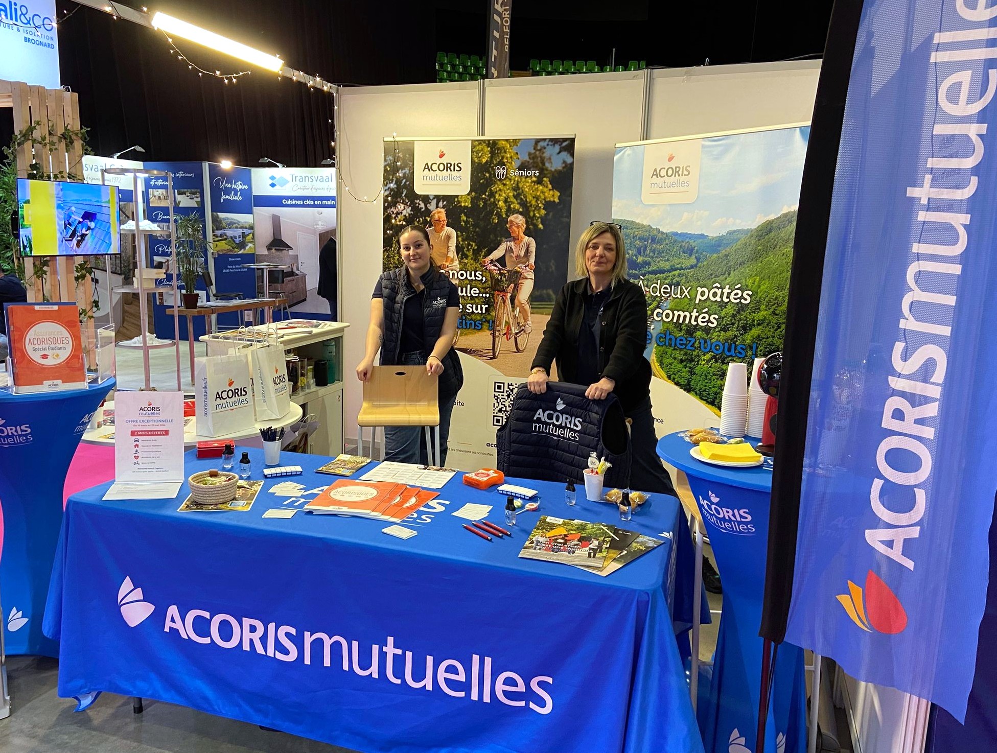 Stand ACORIS Mutuelles sur un salon, avec une table habillée aux couleurs de la mutuelle, des supports de communication et deux personnes accueillant les visiteurs devant des kakémonos illustrant les engagements santé et prévention.
