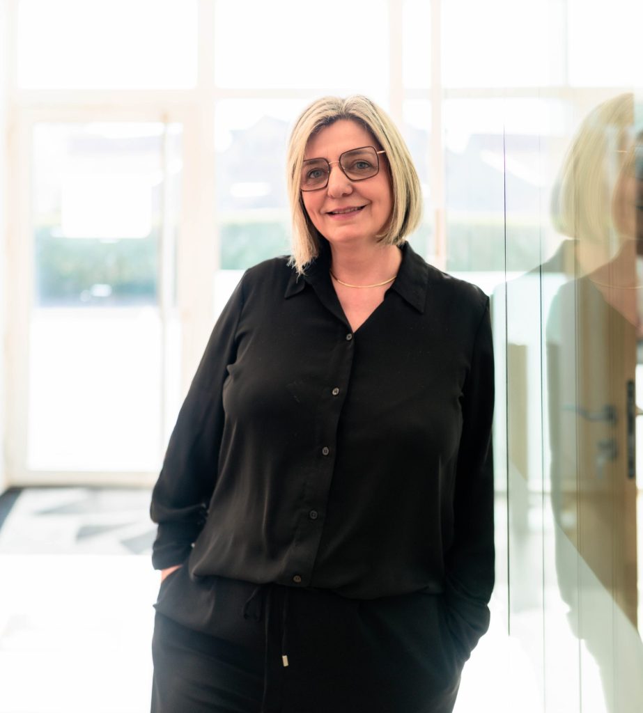 Portrait d’une femme souriante, portant une tenue noire et des lunettes, debout dans un espace lumineux avec une paroi en verre réfléchissante. 