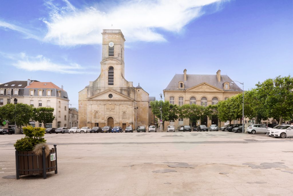 Vue d’une grande place bordée de bâtiments historiques, dont une église en pierre et un édifice à l’architecture classique, avec des voitures garées le long des arbres.