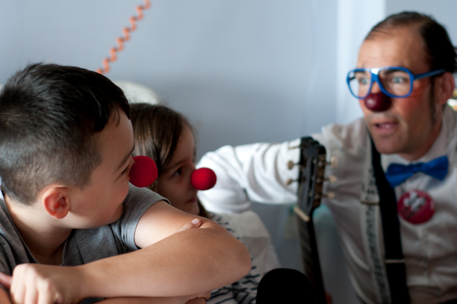 Deux enfants portant des nez de clown regardent un clown musicien, assis près d’eux, dans une pièce lumineuse. 
