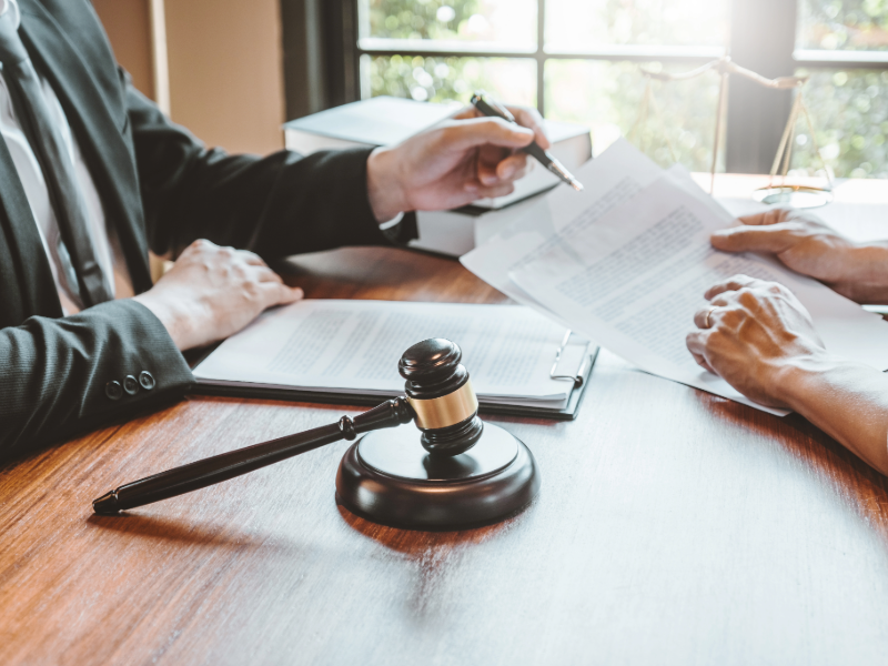 Un marteau de juge posé sur un bureau en bois, au premier plan, tandis que deux personnes assises échangent des documents juridiques et les examinent ensemble.