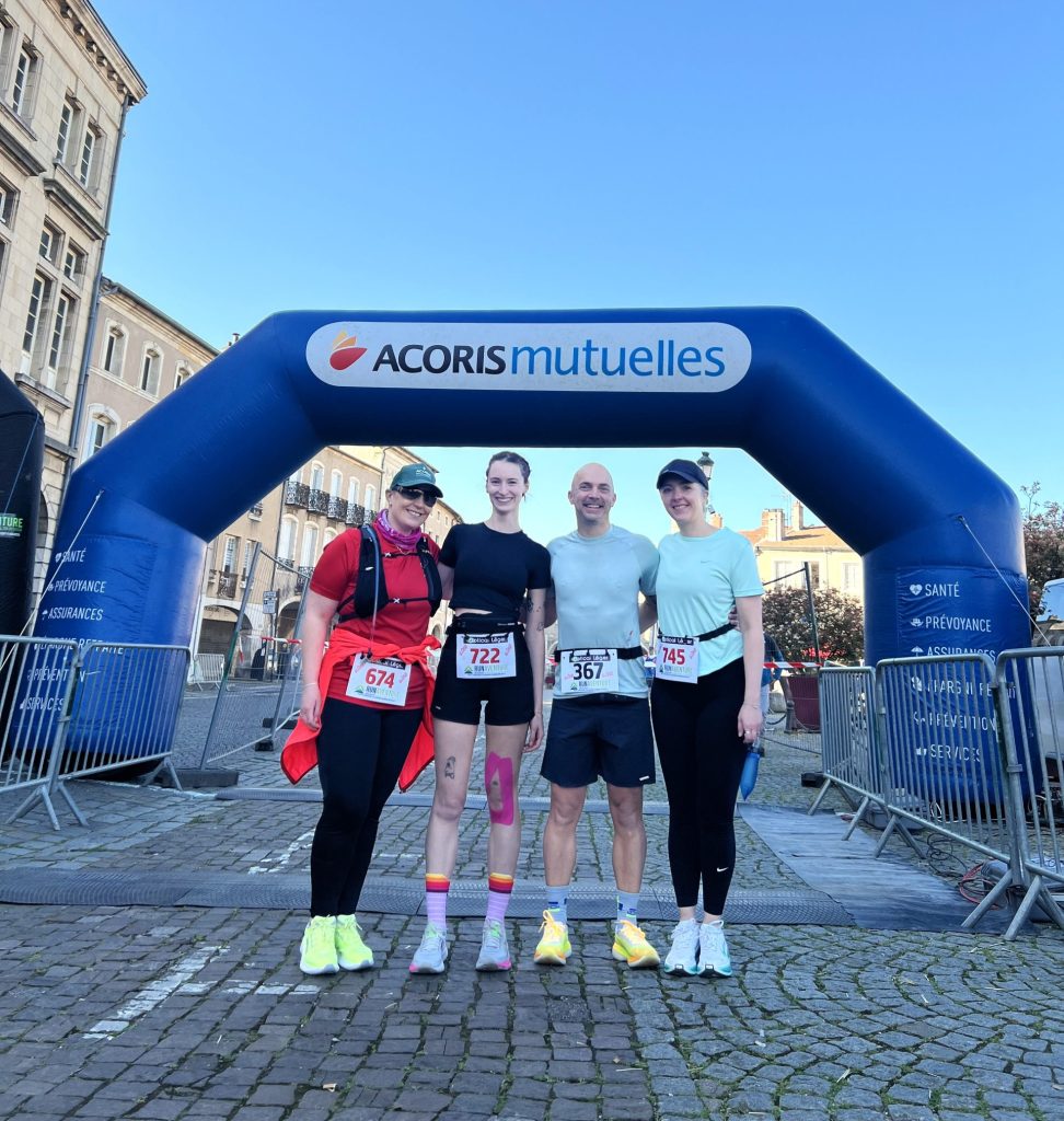 Quatre participants portent des tenues de course et des dossards, posant sous une arche gonflable « ACORIS mutuelles » sur une place pavée, à l’arrivée d’un événement sportif en ville, sous un ciel dégagé.