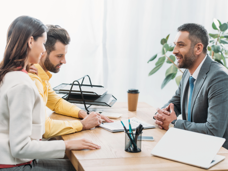 Trois personnes sont assises autour d’une table lors d’une réunion professionnelle, échangeant des documents et prenant des notes dans un espace de bureau lumineux. 