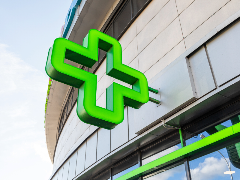 Croix verte lumineuse de pharmacie fixée à la façade d’un bâtiment moderne, avec des baies vitrées et un ciel légèrement nuageux en arrière-plan.
