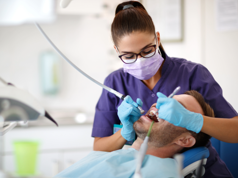 Une dentiste portant un masque, des gants et des lunettes de protection examine la bouche d’un patient allongé sur un fauteuil, à l’aide d’instruments dentaires.
