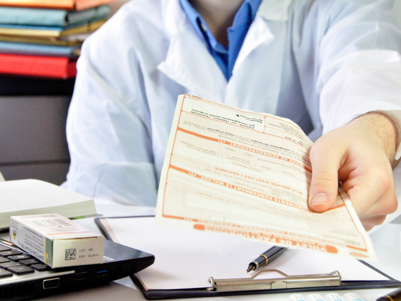Une personne en blouse blanche tend un document médical au‑dessus d’un bureau, avec un stylo, un carnet et une boîte de médicament visibles au premier plan.