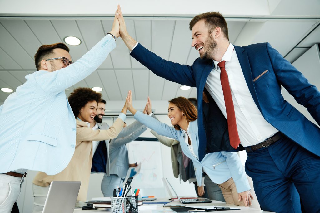 Groupe de personnes en tenue professionnelle se donnant un high five autour d’une table de réunion, dans un espace de bureau lumineux avec ordinateur portable, documents et tableau en arrière-plan.