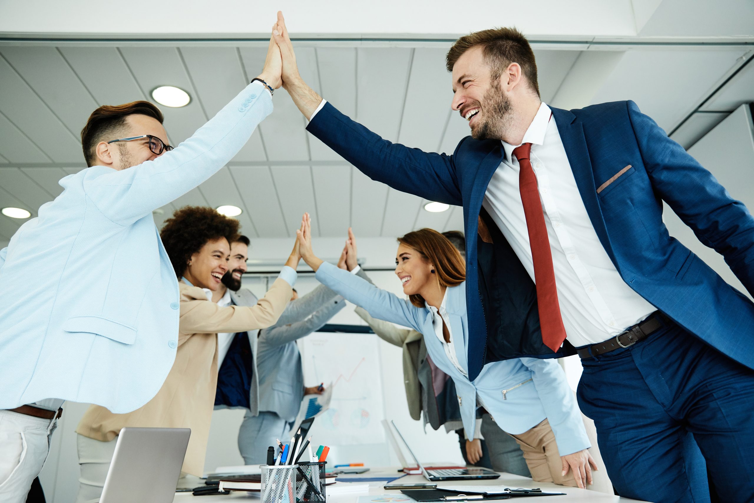 Groupe de personnes en tenue professionnelle se donnant un high five autour d’une table de réunion, dans un espace de bureau lumineux avec ordinateur portable, documents et tableau en arrière-plan.