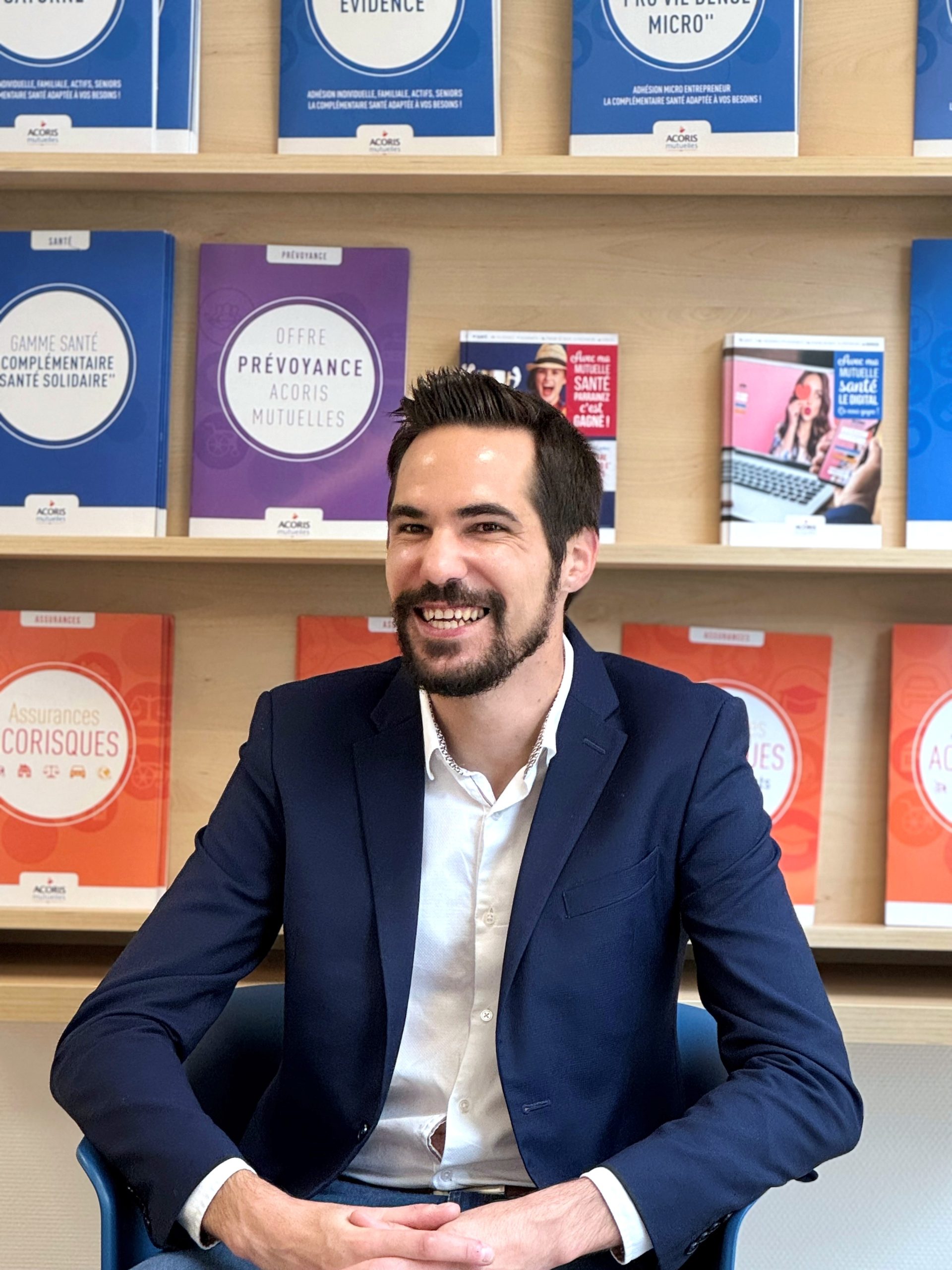 Personne assise sur un fauteuil, portant une veste bleu foncé et une chemise claire, devant une étagère en bois contenant plusieurs brochures d’assurances et de mutuelles aux couleurs bleu, violet et orange.