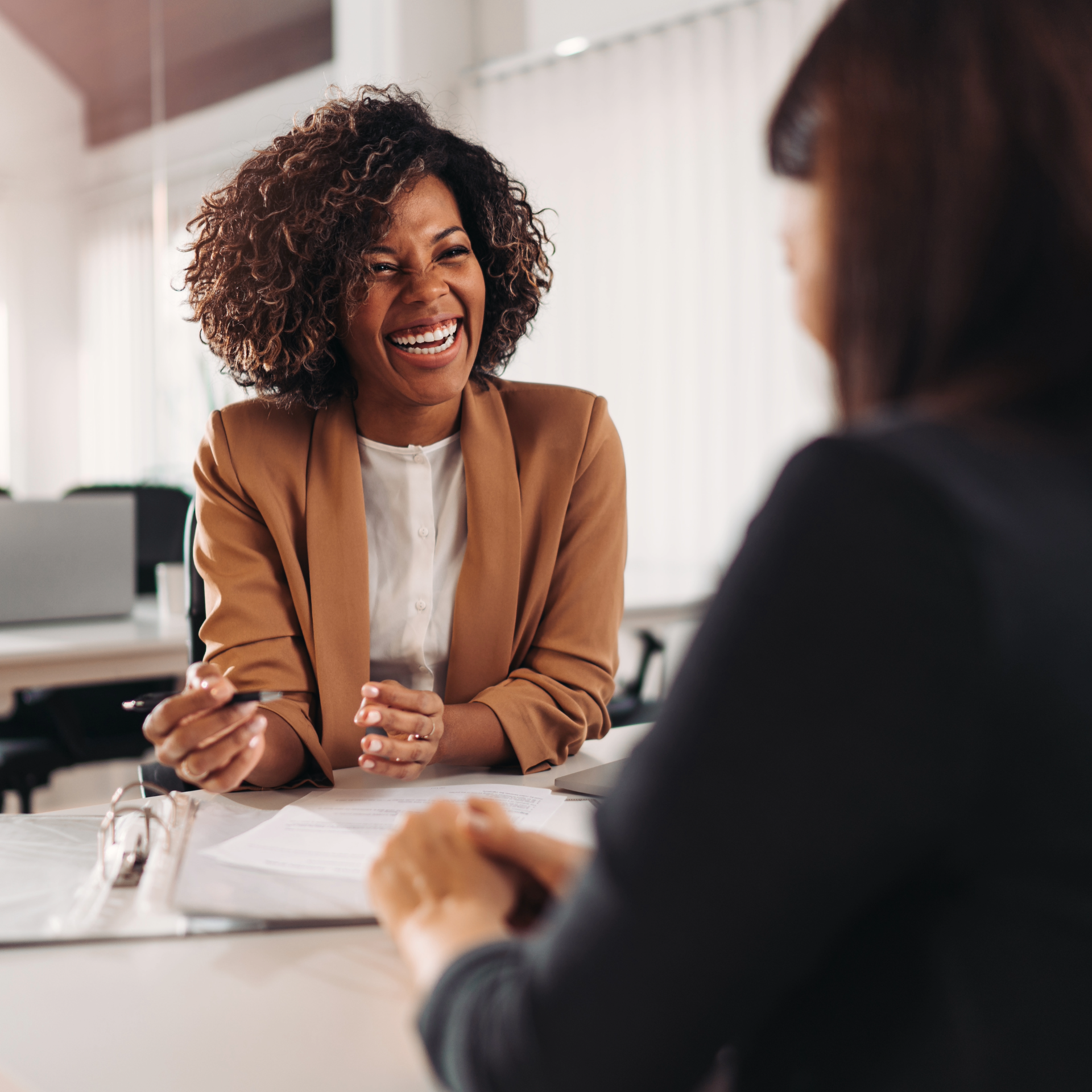 Personne assise à un bureau en train de discuter avec une autre personne hors champ, tenant un stylo et faisant face à des documents posés sur la table, dans un environnement de bureau lumineux.