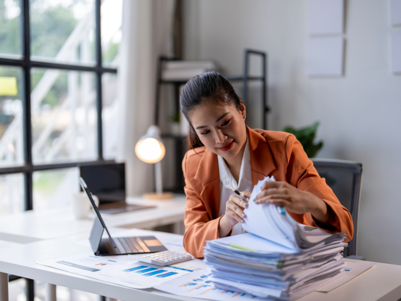 Personne travaillant à un bureau dans un environnement de bureau moderne, consultant et classant des documents papier à côté d’un ordinateur portable et de graphiques financiers.