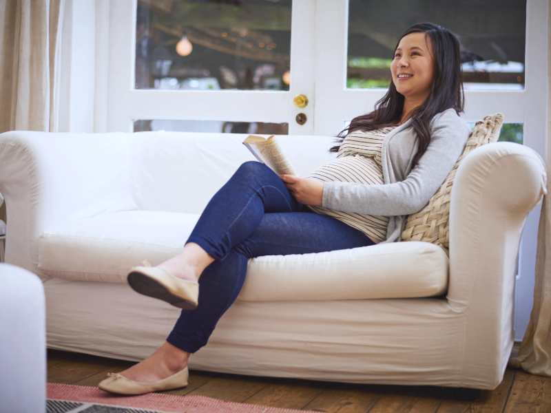 Personne enceinte assise sur un canapé clair, lisant un livre dans un intérieur lumineux, avec une atmosphère confortable et détendue.