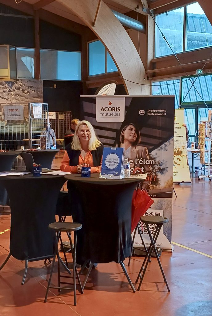 Stand ACORIS Mutuelles lors d’un événement, avec une personne assise à une table haute entourée de supports de communication et de stands professionnels en arrière-plan.