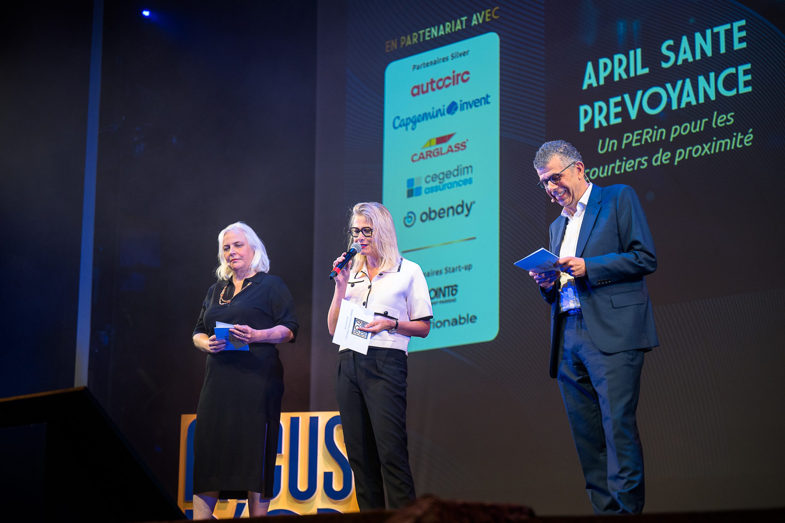 Trois personnes sur une scène éclairée lors d’un événement professionnel, tenant des cartes et un micro, devant un écran affichant “Argus d’Or”, “April Santé Prévoyance” et des logos de partenaires.