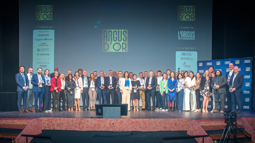 Grand groupe de participants réunis sur scène lors de la cérémonie Argus d’Or, certains tenant des trophées, devant un écran affichant le logo “Argus d’Or” et des partenaires.