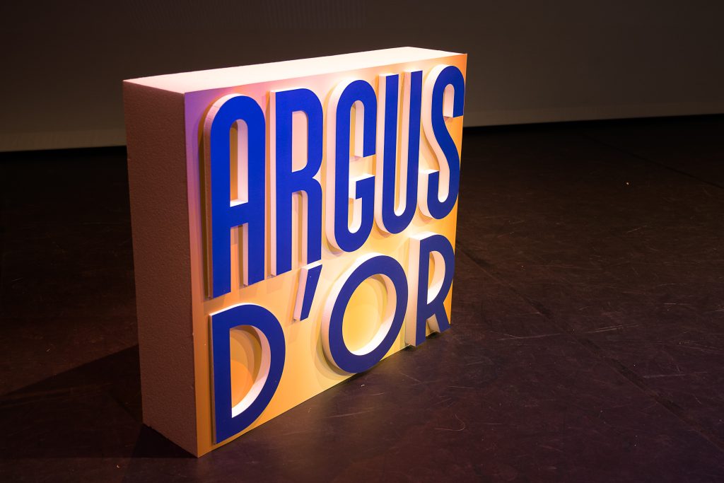 Bloc lumineux posé au sol avec l’inscription en grandes lettres bleues “ARGUS D’OR”, éclairé dans un environnement de scène événementielle.