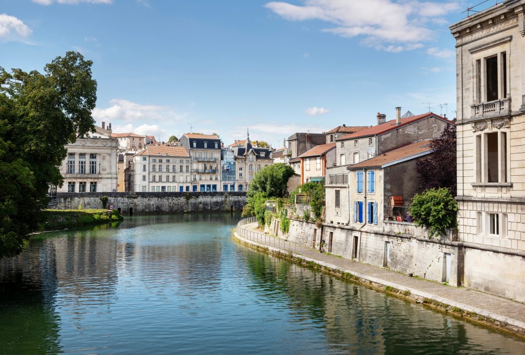 Rivière paisible à Verdun bordée de maisons en pierre aux toits rouges, longeant un quai arboré sous un ciel bleu clair.