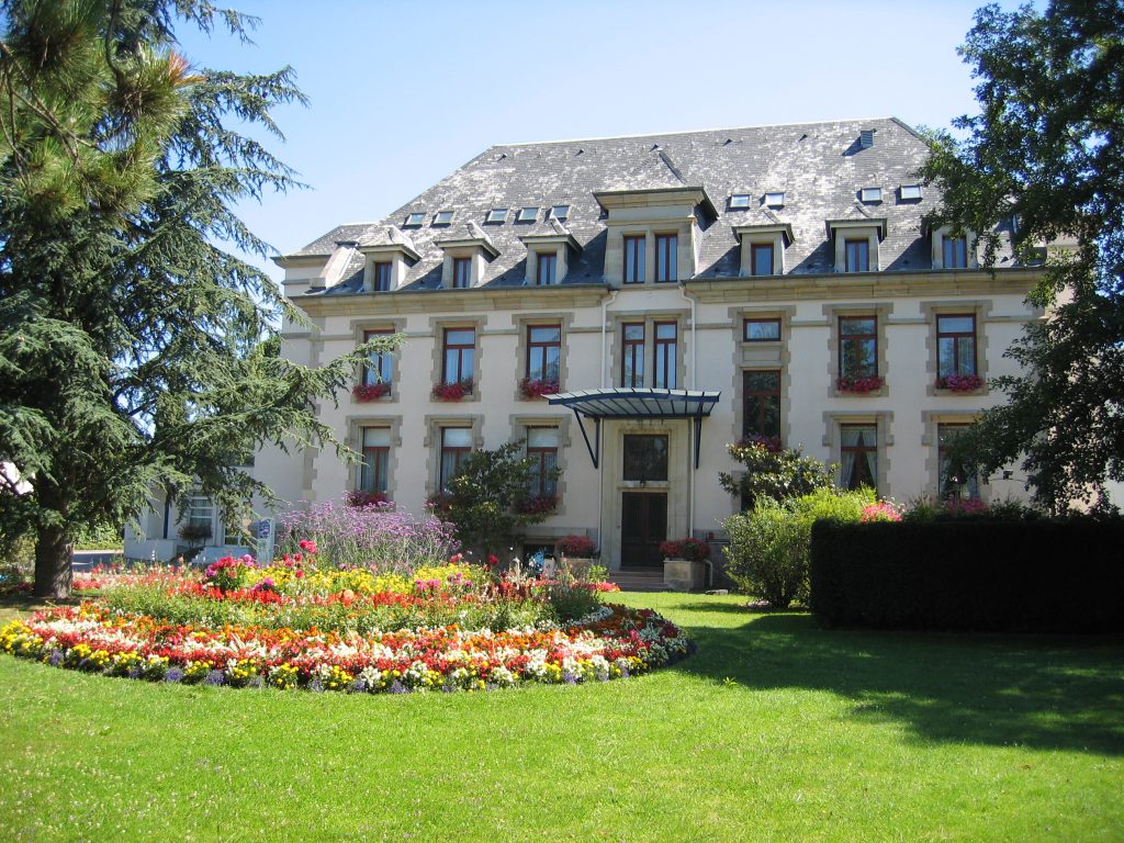 Bâtiment élégant de style classique, avec toiture mansardée et fenêtres ornées de fleurs, entouré d’un jardin bien entretenu avec une pelouse verdoyante et un massif de fleurs colorées au premier plan, sous un ciel bleu dégagé.