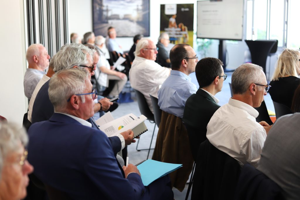 Participants assis lors d’une réunion ou assemblée, certains tenant des documents ou prenant des notes, dans une salle de conférence lumineuse.