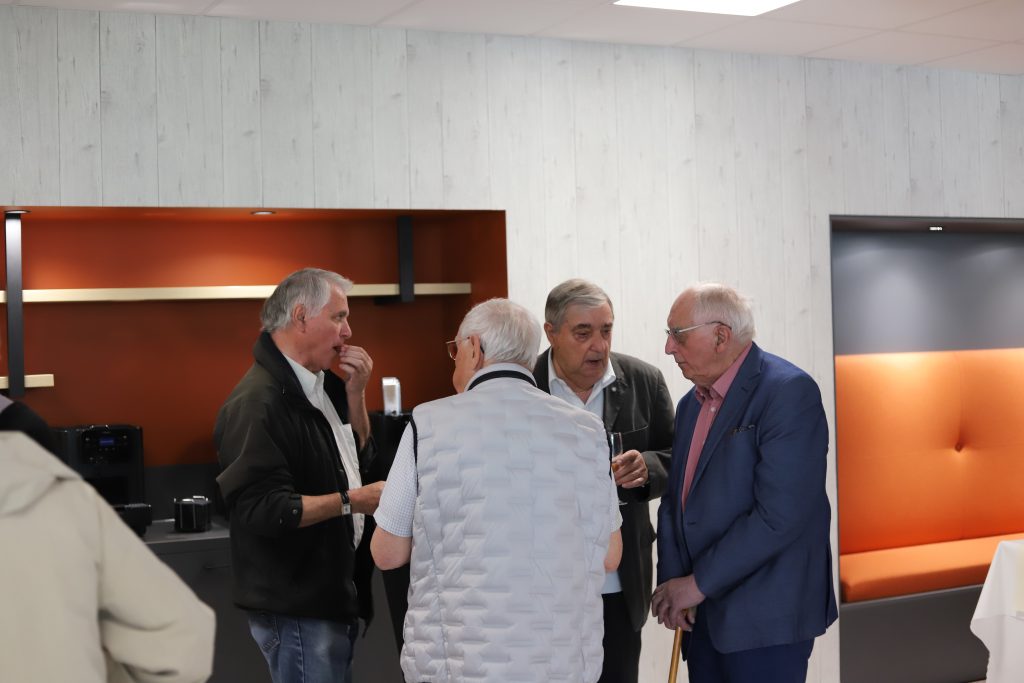 Groupe de participants discutant autour d’un espace de pause, certains tenant des verres, dans un environnement intérieur moderne avec mur décoratif et coin salon.