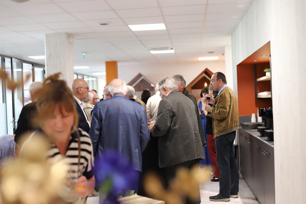 Participants réunis lors d’un moment convivial autour d’un buffet dans un espace intérieur moderne, discutant en petits groupes, certains tenant des verres près d’un comptoir avec machine à café.