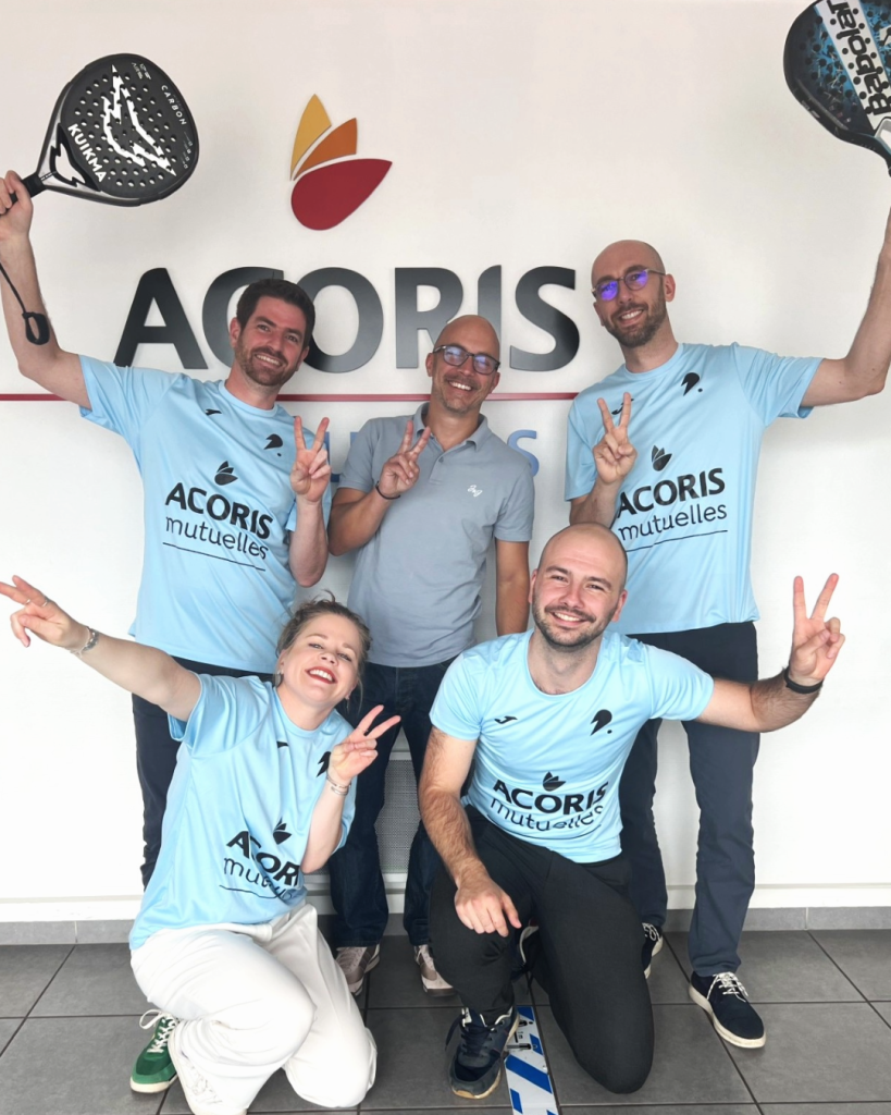 Cinq personnes en t-shirts « ACORIS mutuelles » posent avec des raquettes de padel devant un mur avec le logo ACORIS, en faisant le signe de victoire.