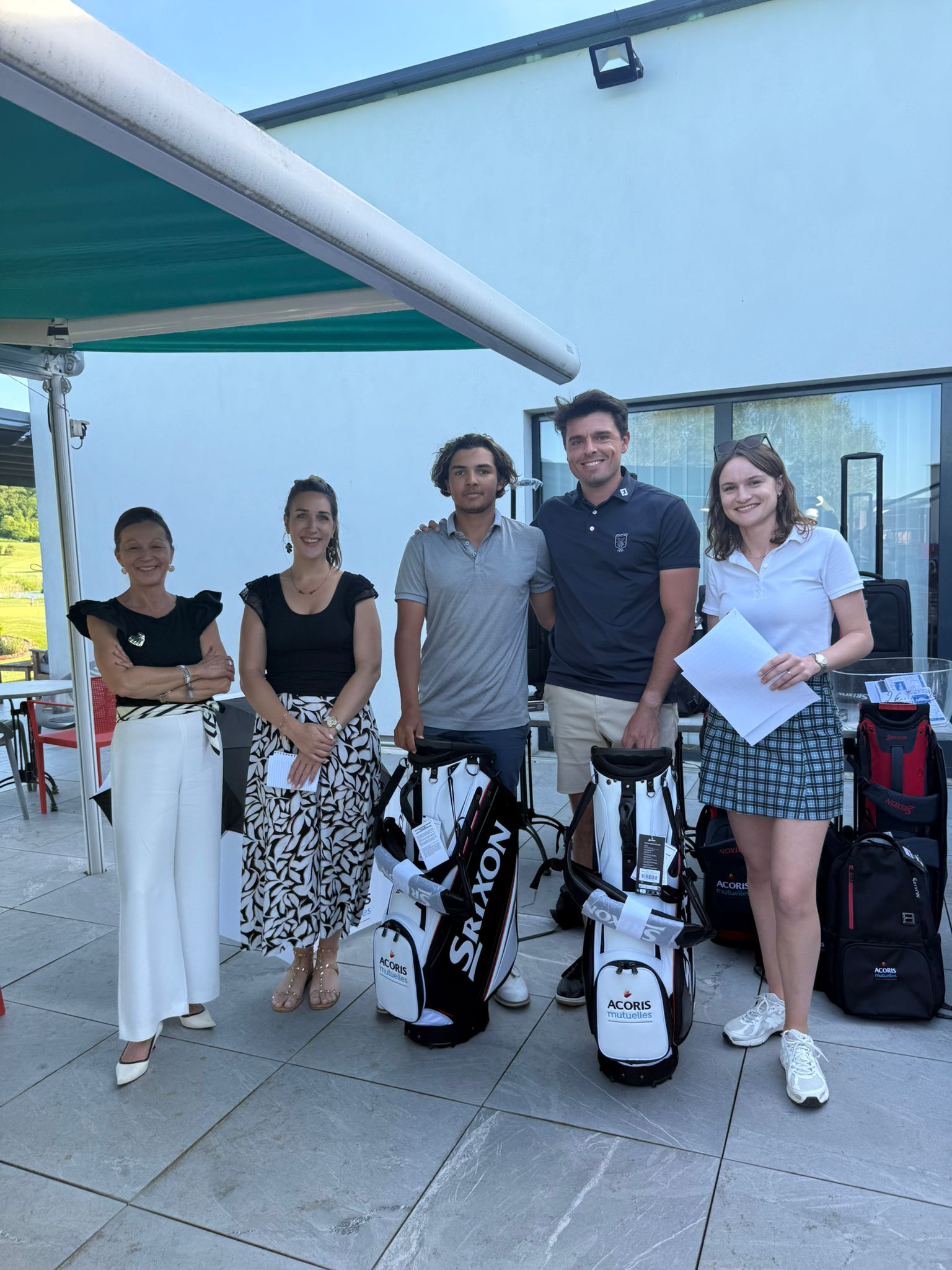 Cinq personnes posant côte à côte sur une terrasse extérieure devant un bâtiment moderne, avec plusieurs sacs de golf et équipements marqués «ACORIS Mutuelles », sous un store vert, avec baies vitrées et paysage verdoyant en arrière-plan.