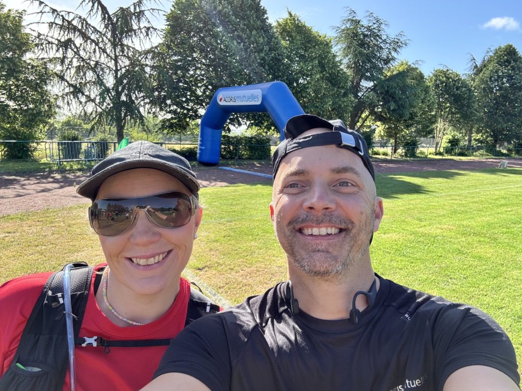 Deux personnes prennent un selfie en extérieur, sur une pelouse arborée par temps ensoleillé. En arrière‑plan, une arche gonflable bleue portant le texte « ACORIS Mutuelles » est installée près d’un chemin, avec des arbres et des barrières métalliques autour.