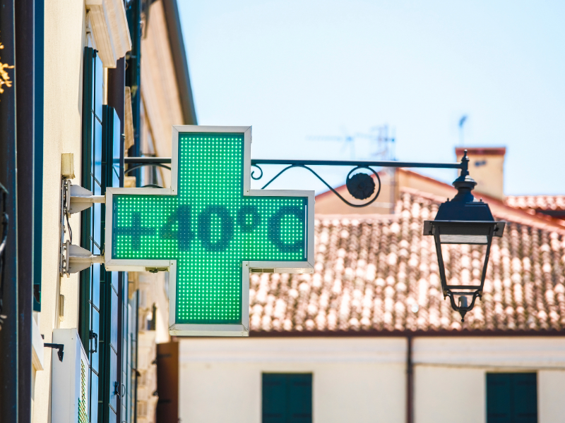 Enseigne lumineuse en forme de croix verte affichant « +40°C », fixée à la façade d’un bâtiment en rue. À côté, un lampadaire noir suspendu et, en arrière-plan, des toits en tuiles et un ciel dégagé.