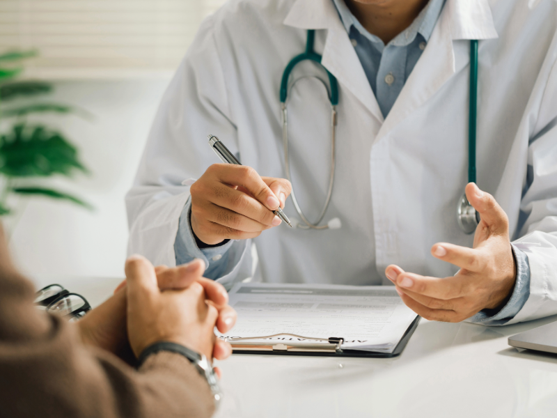 Personne portant une blouse médicale avec un stéthoscope autour du cou, assise à un bureau, échangeant avec une autre personne. Une main tient un stylo au-dessus d’un document posé sur une planchette, tandis que l’autre main est ouverte en geste d’explication. Au premier plan, les mains de l’interlocuteur sont visibles, posées l’une contre l’autre. L’environnement est un cabinet médical lumineux avec un fond légèrement flou.