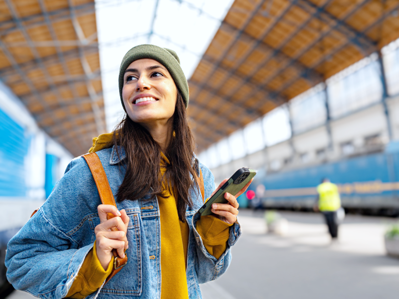Personne debout sur un quai de gare moderne, tenant un smartphone dans une main et portant un sac à dos. La personne porte un bonnet vert, une veste en jean et un sweat à capuche jaune. Un train bleu est visible à l’arrière-plan, sous une grande verrière métallique offrant une lumière naturelle.