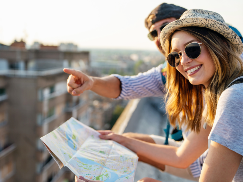 Deux personnes surplombant une ville depuis un point élevé consultent une carte papier. Une personne au premier plan porte un chapeau clair et des lunettes de soleil rondes, tient la carte et se tourne vers l’appareil photo, tandis qu’une autre personne à l’arrière-plan, également avec des lunettes de soleil, pointe du doigt vers l’horizon. Des bâtiments urbains et un ciel lumineux sont visibles en arrière-plan.