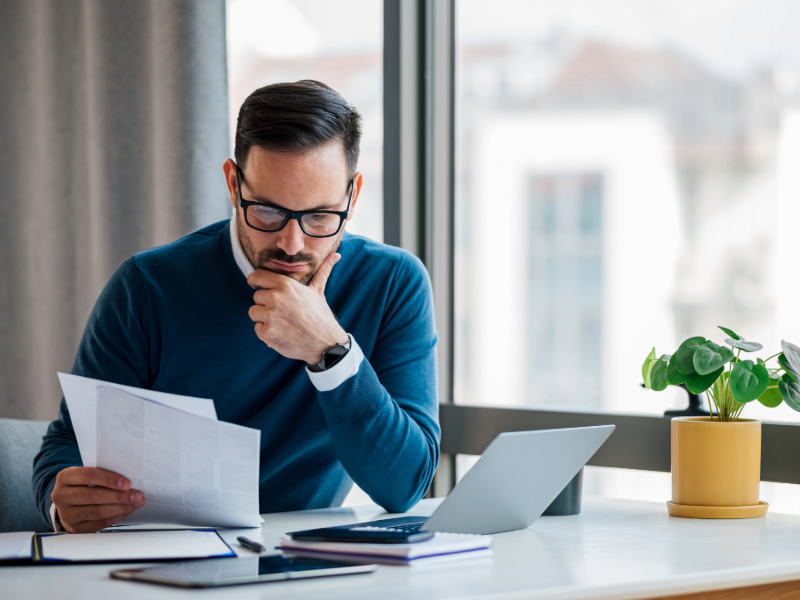Personne portant des lunettes, assise à un bureau près d’une fenêtre, examinant attentivement plusieurs documents papier tout en tenant un stylo. Un ordinateur portable ouvert, un smartphone et un carnet sont posés sur la table, avec une plante verte en pot à proximité, dans un environnement lumineux et épuré.