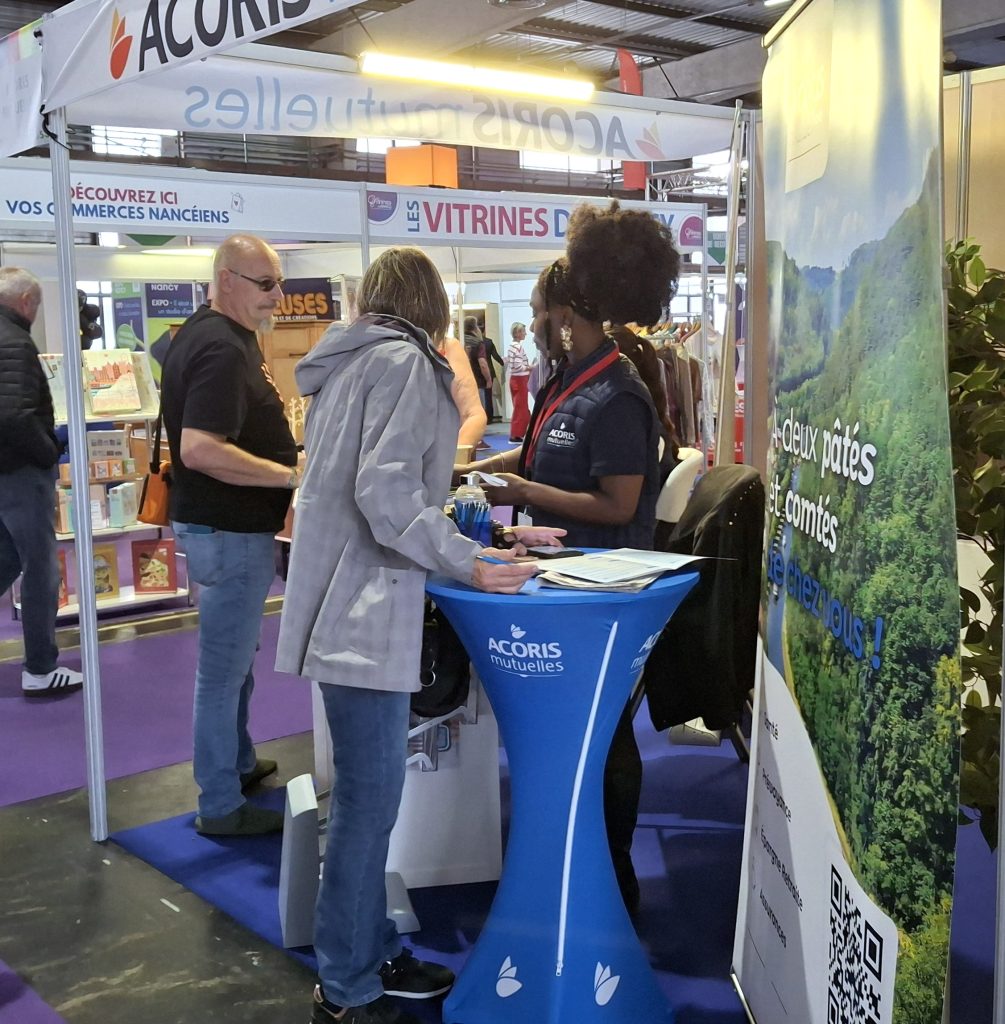 Stand ACORIS Mutuelles sur un salon avec un conseiller échangeant avec des visiteurs autour d’un espace d’information sur la santé et la prévoyance, dans un hall d’exposition accueillant des exposants locaux.