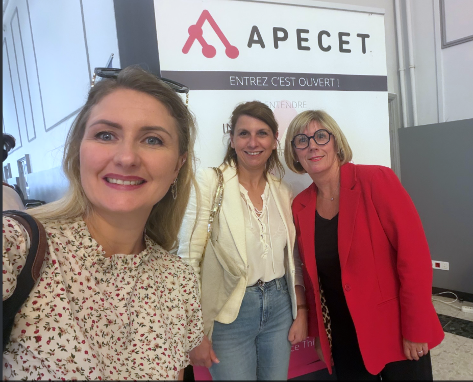 Trois personnes posent pour un selfie devant un stand APECET avec le slogan “Entrez c’est ouvert !”. Le stand présente un visuel sobre avec un logo rouge et noir. L'environnement est un espace intérieur de type événement professionnel avec murs clairs et signalétique.