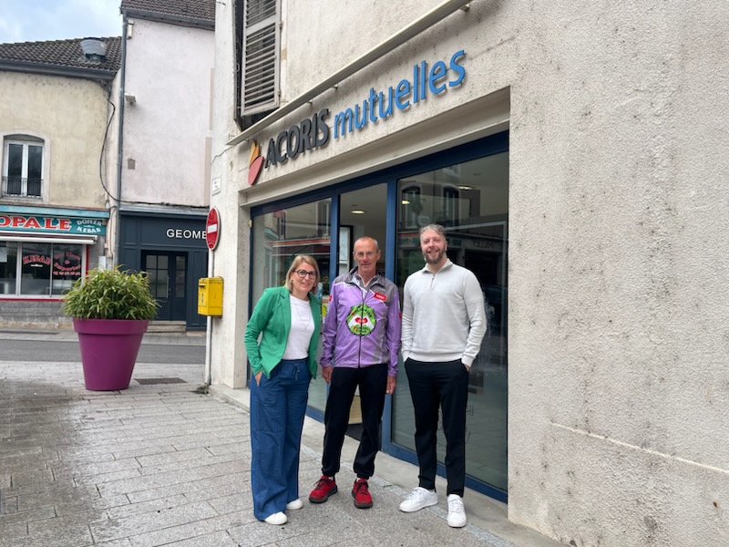 Trois personnes posent devant une agence ACORIS Mutuelles située en centre-ville. Au centre, une personne porte une veste de cyclisme violette ornée d’un logo, tandis que les deux autres sont vêtues de tenues de ville. L’enseigne « Acoris mutuelles » est visible au-dessus de la vitrine et de l’entrée de l’agence. La scène se déroule sur un trottoir bordé de commerces, avec un grand pot de fleurs violet à proximité. Cette photo illustre une rencontre entre un participant à un événement cycliste et des représentants de l’agence locale.