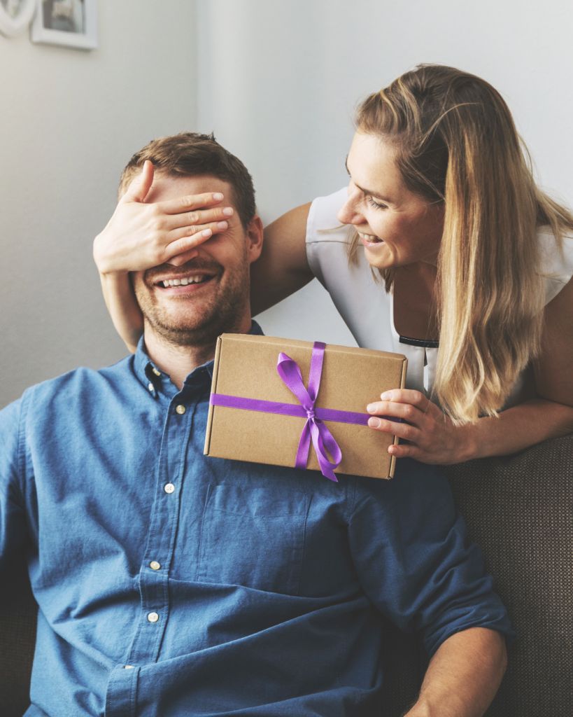 Deux personnes sont assises sur un canapé dans un intérieur lumineux. L’une est assise au premier plan et tient un coffret cadeau en carton décoré d’un ruban violet. L’autre se penche au-dessus de son épaule et place une main devant ses yeux. Le cadeau est présenté au centre de l’image. Un mur clair constitue l’arrière-plan, tandis que la scène met en avant un moment de surprise autour de l’offre d’un présent.