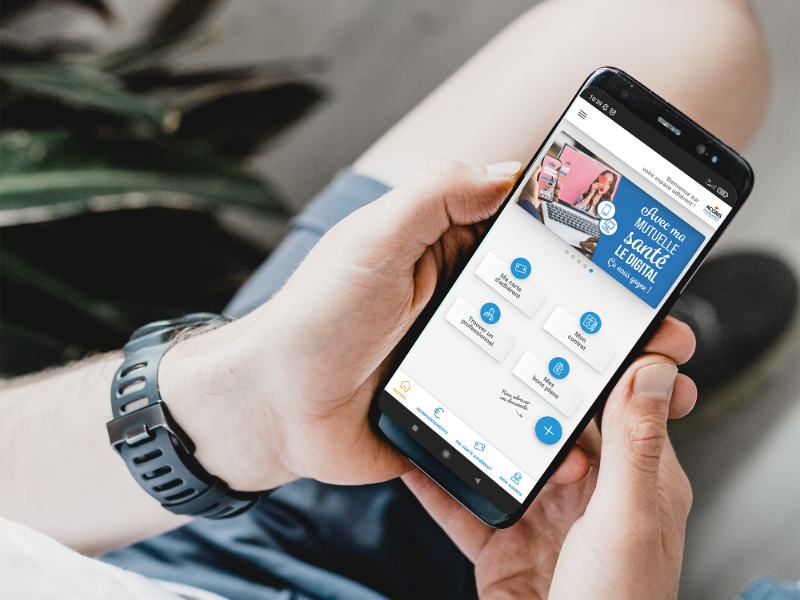 Gros plan sur une personne tenant un smartphone dans ses mains. L’écran affiche l’interface d’une mutuelle avec plusieurs raccourcis de services et une bannière promotionnelle bleue. L’arrière-plan est flou, avec un bracelet de montre visible au poignet et une plante verte en arrière-plan.