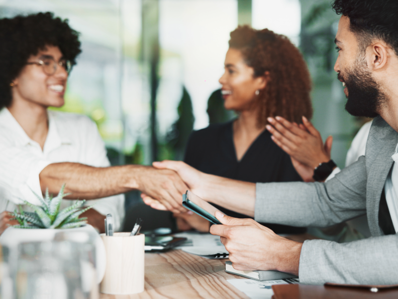 Trois personnes sont réunies autour d’une table de réunion dans un espace de travail lumineux. Au premier plan, deux personnes se serrent la main au-dessus de documents et d’un smartphone, tandis qu’une troisième personne est visible en arrière-plan. Plusieurs objets de bureau, dont un carnet, un stylo et une plante en pot, sont disposés sur la table. L’image illustre un échange professionnel, un accord ou une collaboration dans un contexte d’entreprise.