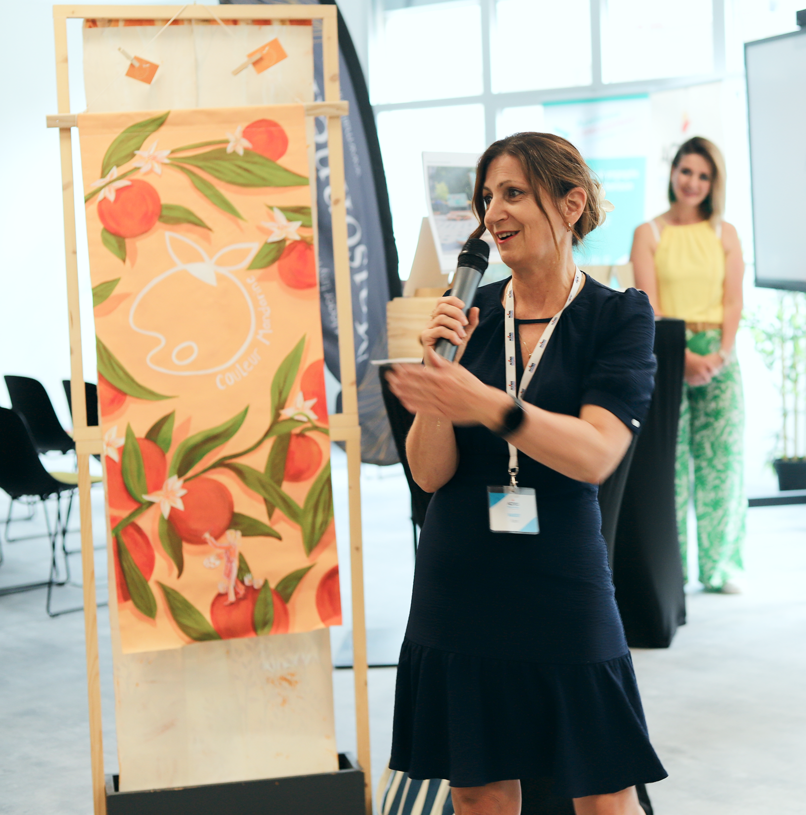 Personne prenant la parole avec un micro lors d’un événement professionnel, à côté d’une œuvre illustrée aux couleurs vives présentée sur un chevalet. Des participants et des stands sont visibles en arrière-plan.