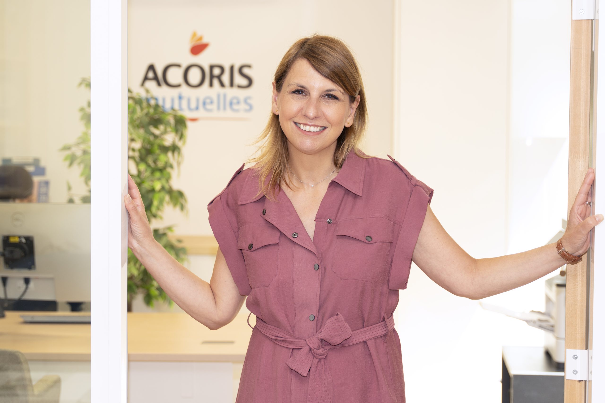 « Personne debout dans l'encadrement d'une porte de bureau, portant une robe rose à ceinture. En arrière-plan, un espace de travail lumineux avec une plante et le logo "ACORIS Mutuelles" visible sur le mur. »