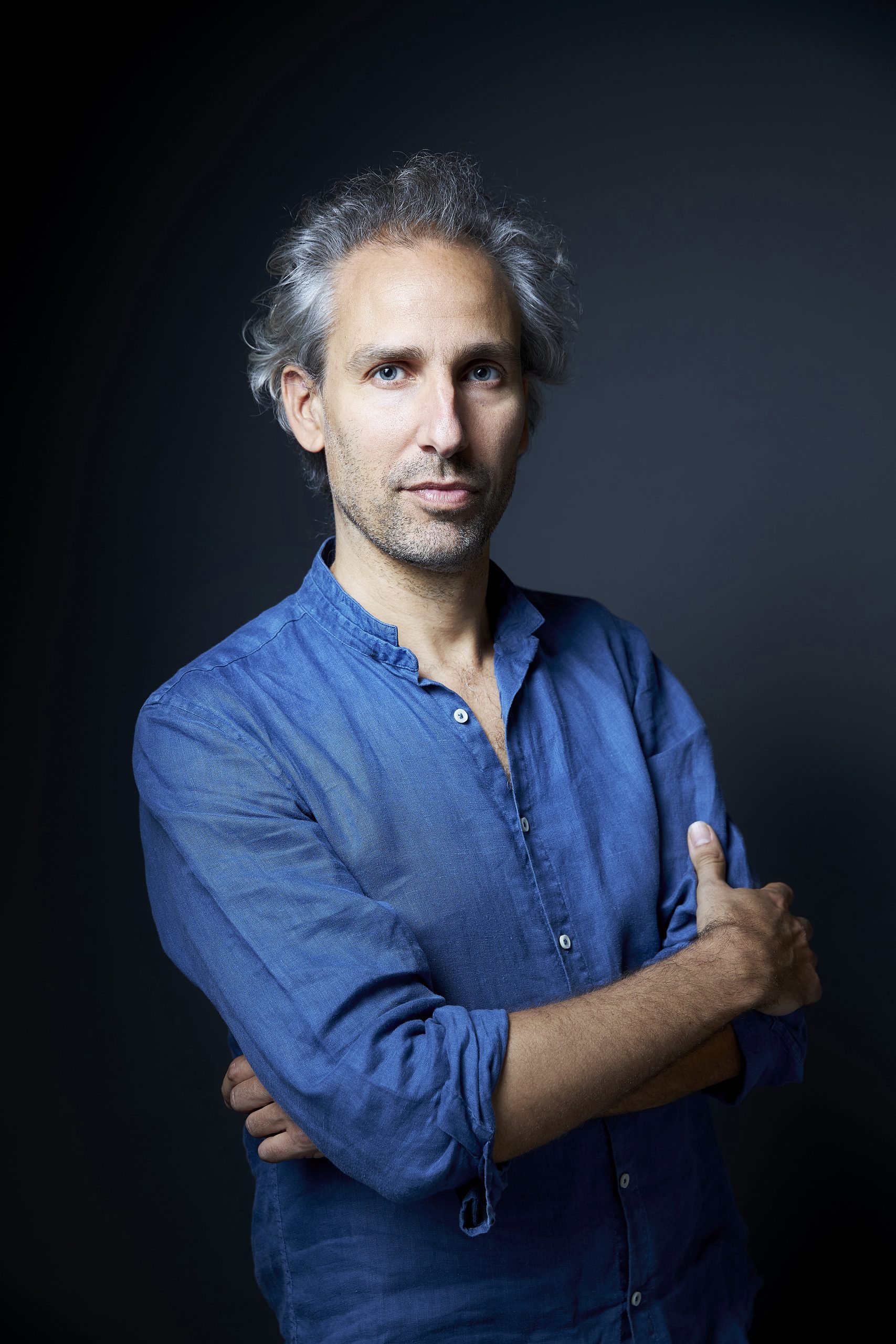 Portrait en studio d’une personne aux cheveux gris portant une chemise bleue à manches retroussées. La personne est photographiée de face, les bras croisés, sur un fond sombre aux nuances de bleu.