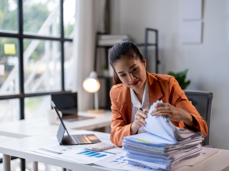 Personne travaillant dans un bureau moderne, consultant et classant une pile de documents administratifs sur un bureau avec tablette numérique et tableaux de données.